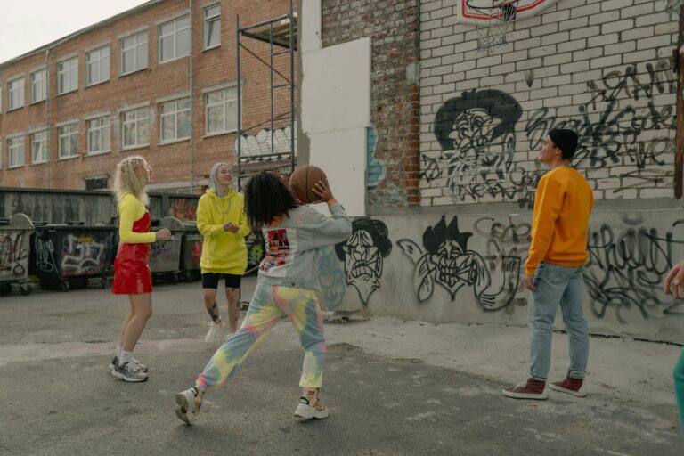 A diverse group plays basketball in an alley with vibrant graffiti in the background.
