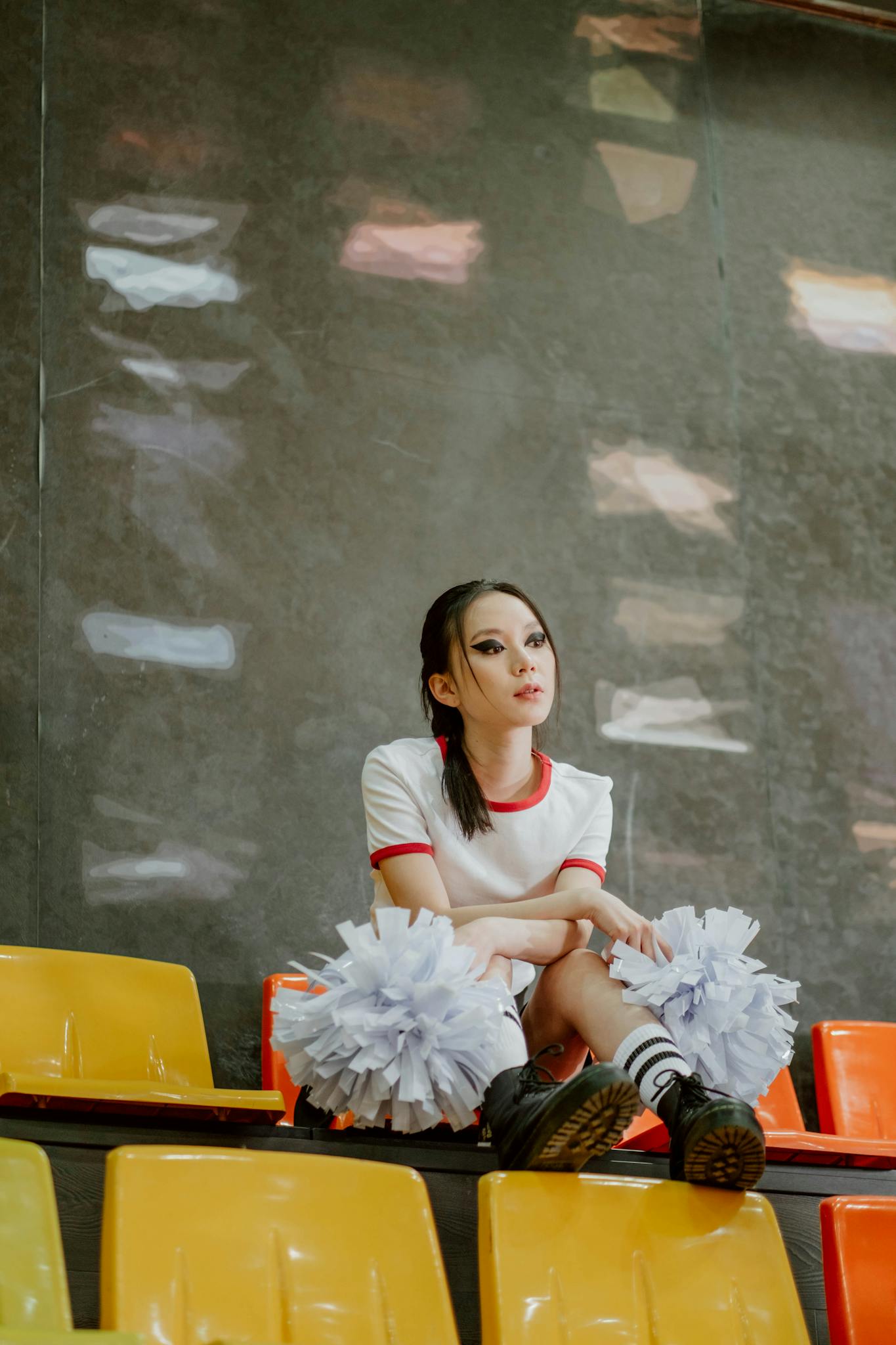 Cheerleader seated on stadium bleachers with pompoms, looking away thoughtfully indoors.