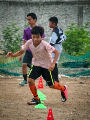 Ethnic kid running through training cones while playing football with team players on field