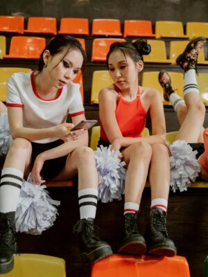 Two female cheerleaders sitting with pompoms, checking a smartphone in a stadium.